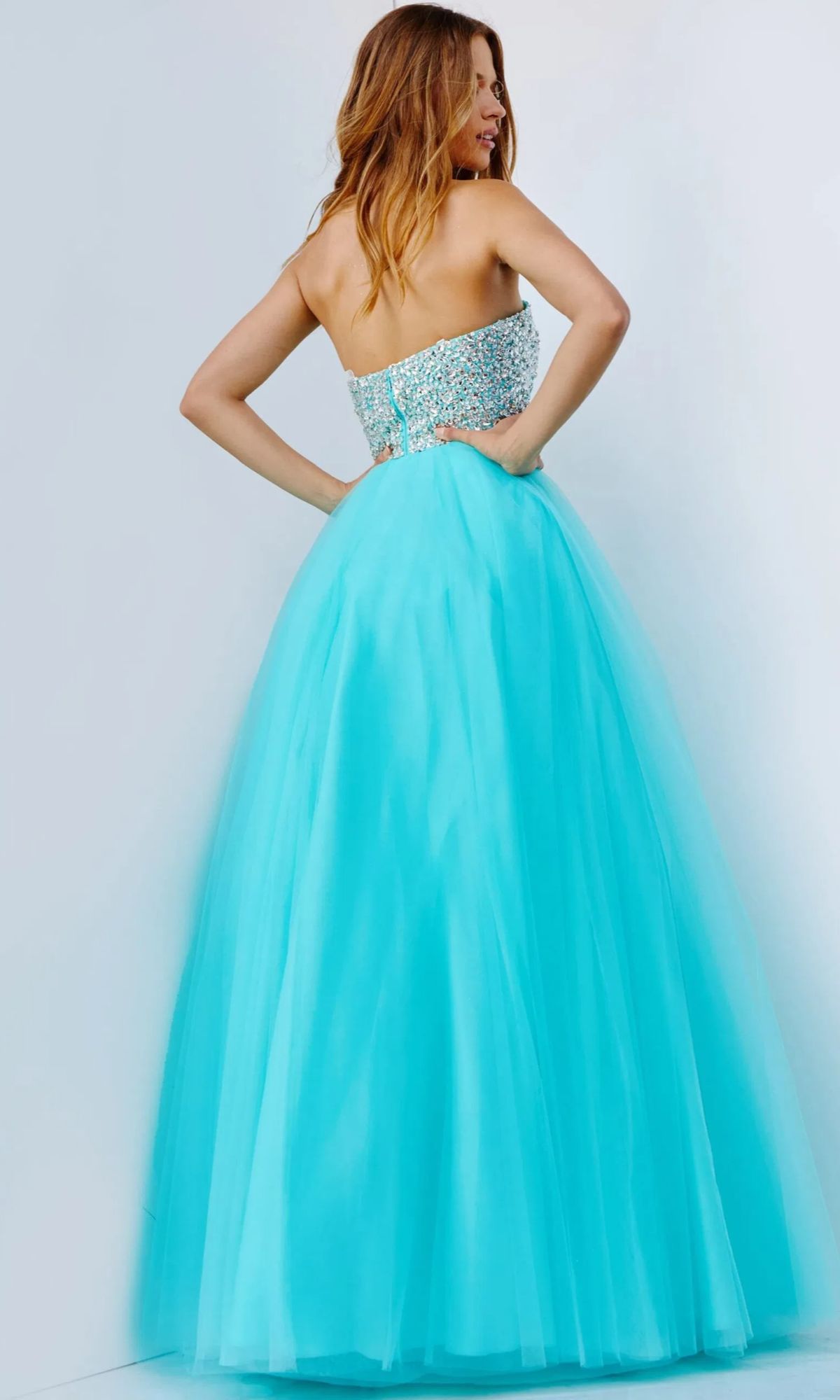 A woman wearing the Long Strapless Ball Gown Prom Dress, Jovani 52131, featuring a beaded bodice, stands facing away with hands on hips against a plain light background.